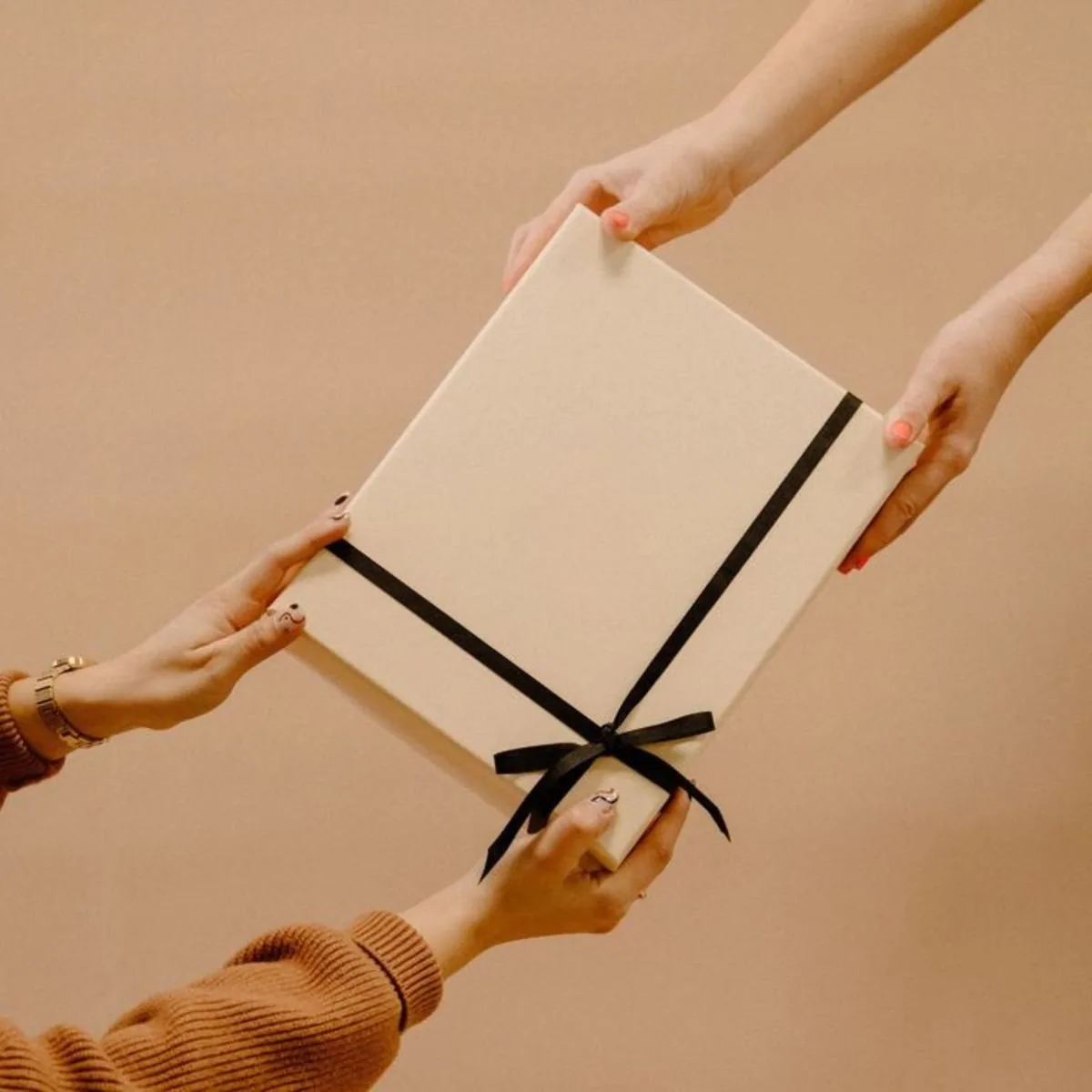 A cream rigid gift box tied with black ribbon being passed from one pair of hands to another
