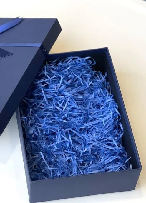A navy rigid lid box open on a white surface with blue paper shred in the base