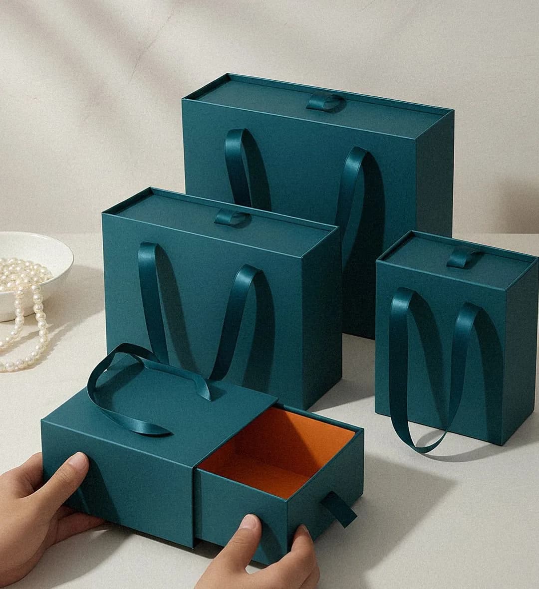 A set of deep teal drawer boxes with ribbon handles, the smallest one being slid open to show an orange lining