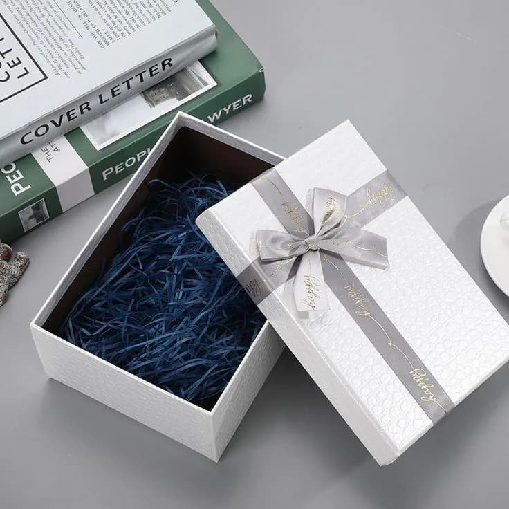 A white embossed lift off lid box open to show navy paper shred, with a grey ribbon tied across the lid